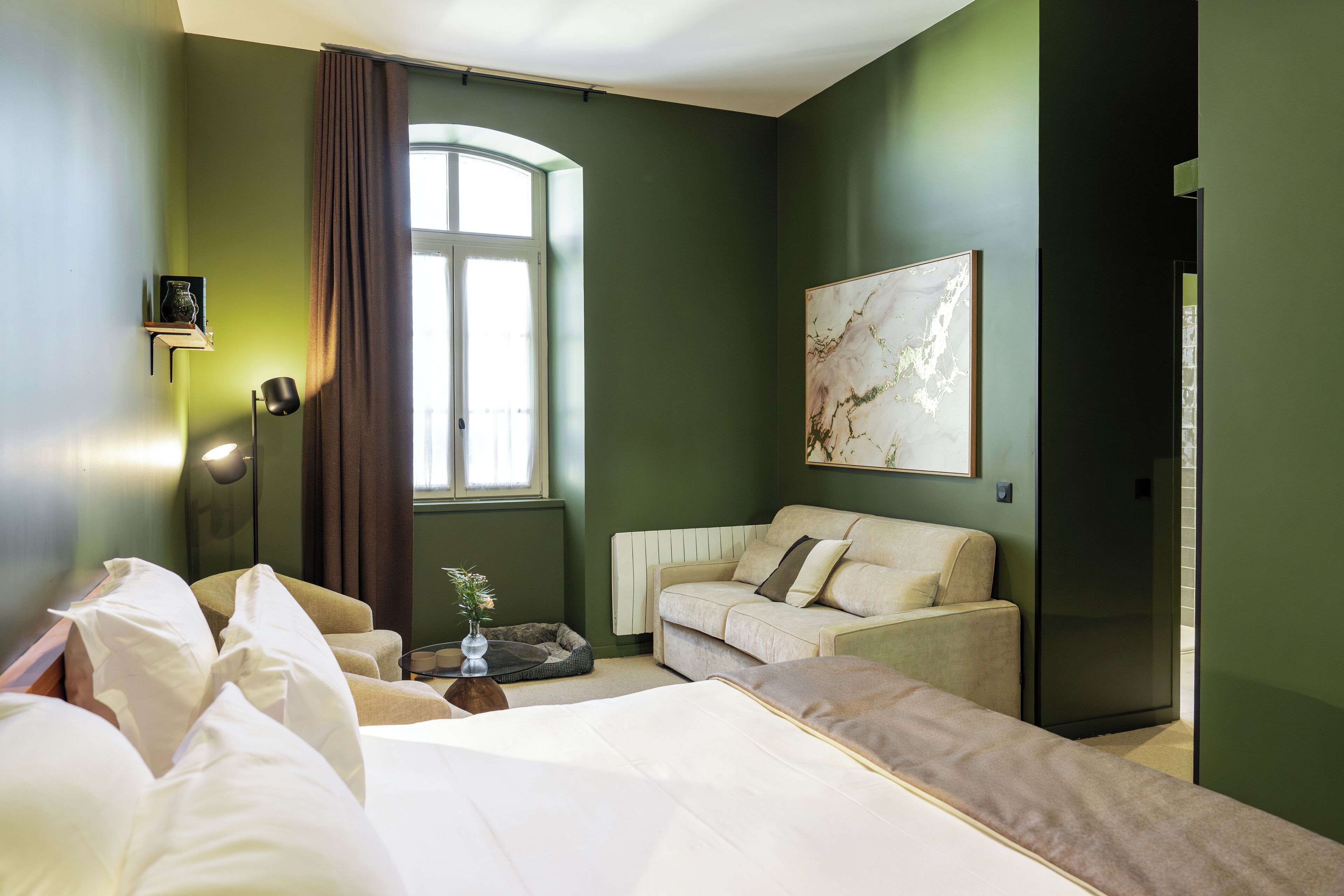 Quarto de hotel com cama, sofá, mesa pequena com um vaso, janela com cortinas e um quadro na parede.