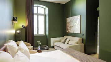 Quarto de hotel com cama, sofá, mesa pequena com um vaso, janela com cortinas e um quadro na parede.