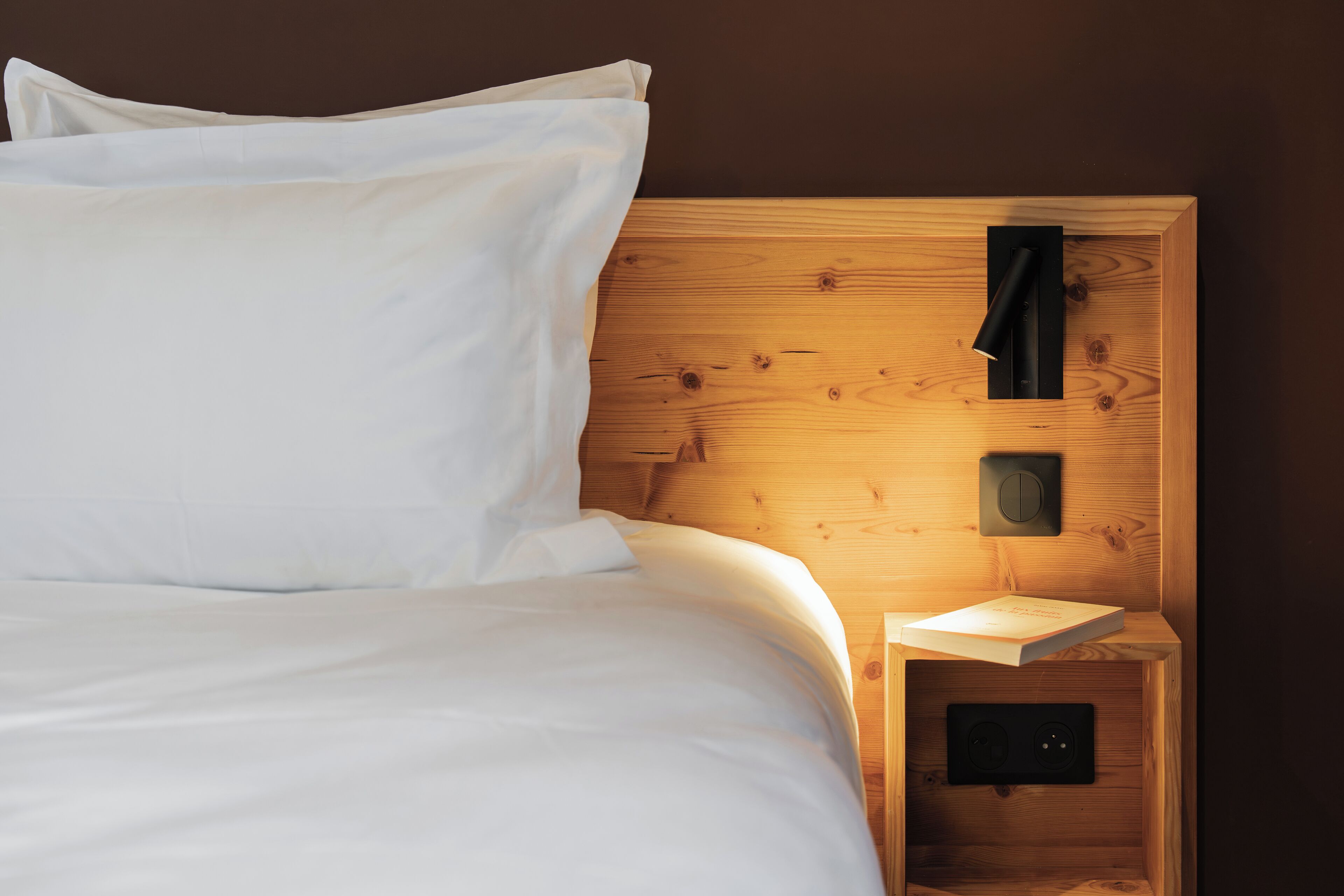 Un lit bien fait, avec du linge de lit blanc, une tête de lit en bois, une table de chevet sur laquelle repose un livre et un luminaire fixé au mur.