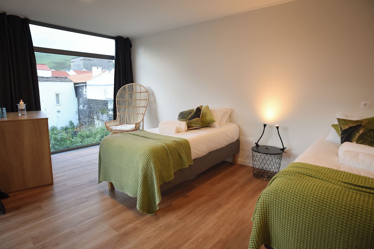 A modern bedroom with a large window, a wooden floor, a bed with green bedding, a chair, and a bedside table.
