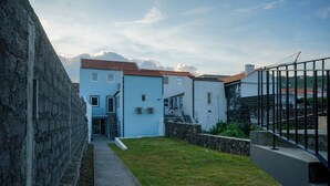 A courtyard with a stone wall, a blue building with a red roof, and a metal fence. - largobispohostel (Horta)