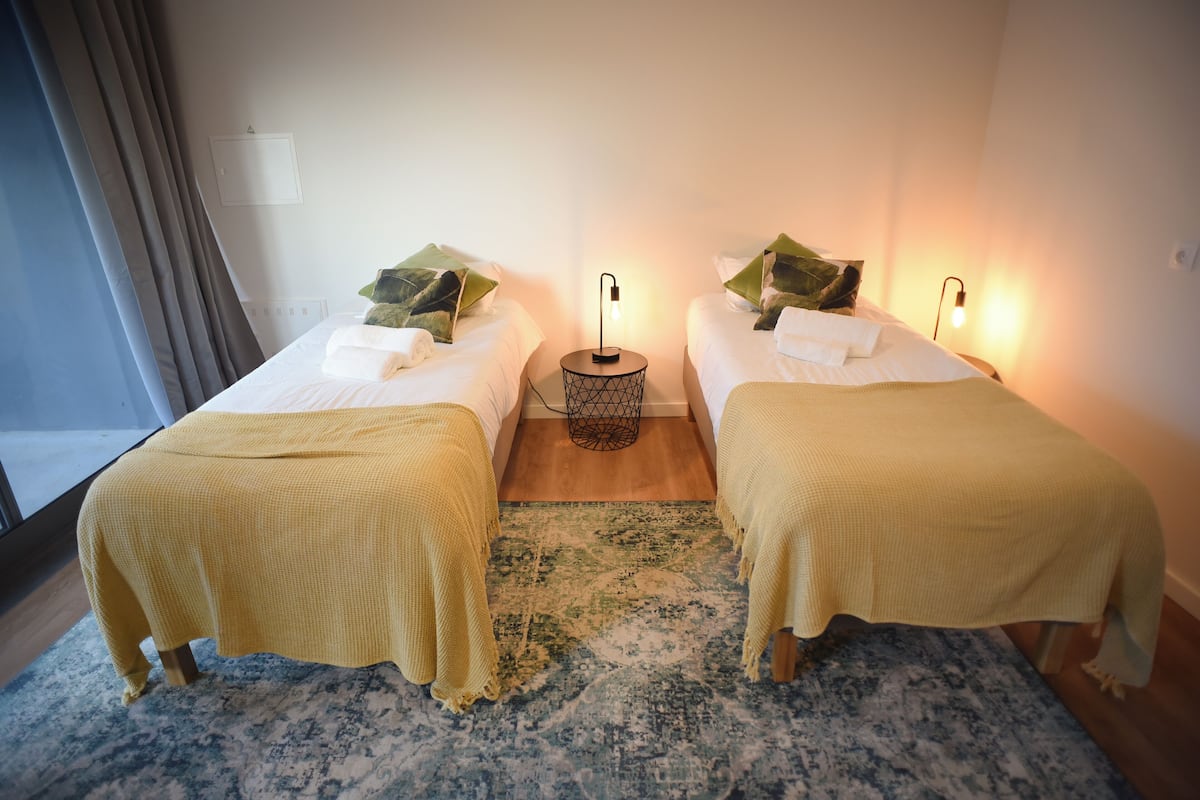 A hotel room with two beds, each with a yellow blanket and green leaves as pillows. There is a small round table with a lamp between the beds.