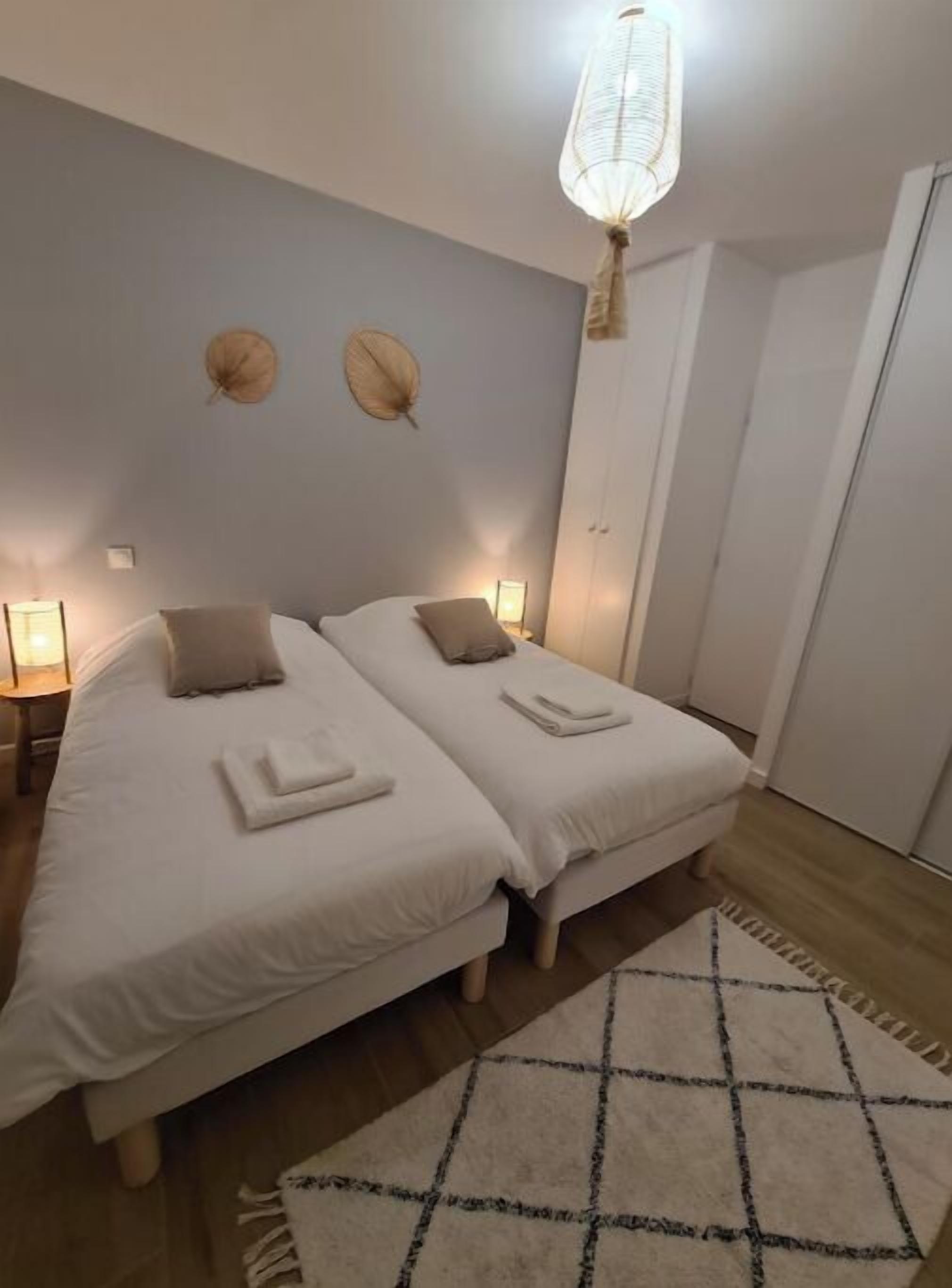 A bedroom with two beds, a nightstand, a lamp, a wall-mounted light fixture, and a door.