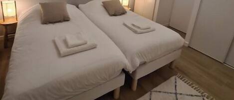 A bedroom with two beds, a nightstand, a lamp, a wall-mounted light fixture, and a door.