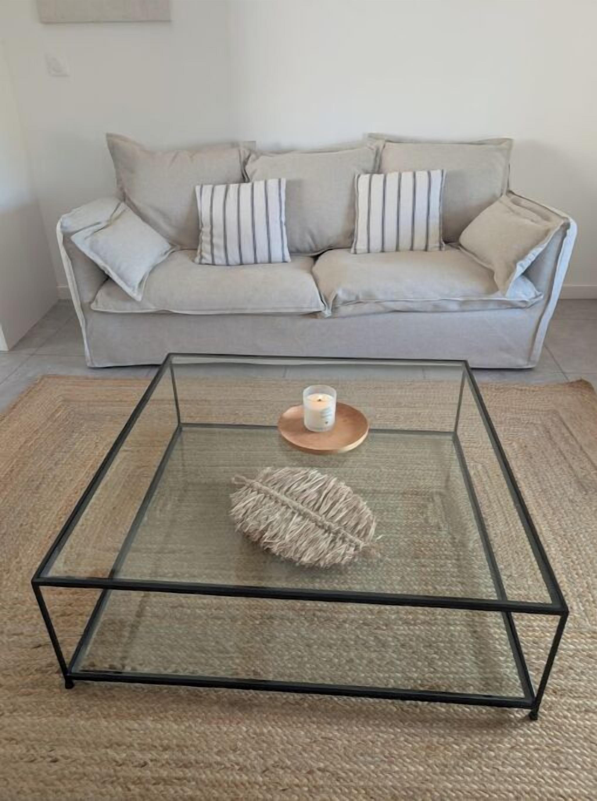 Ein Glastisch mit gewebter Unterlage und einer Kerze auf einem Holzuntersetzer, vor einer hellgrauen Couch mit gestreiften Kissen platziert.