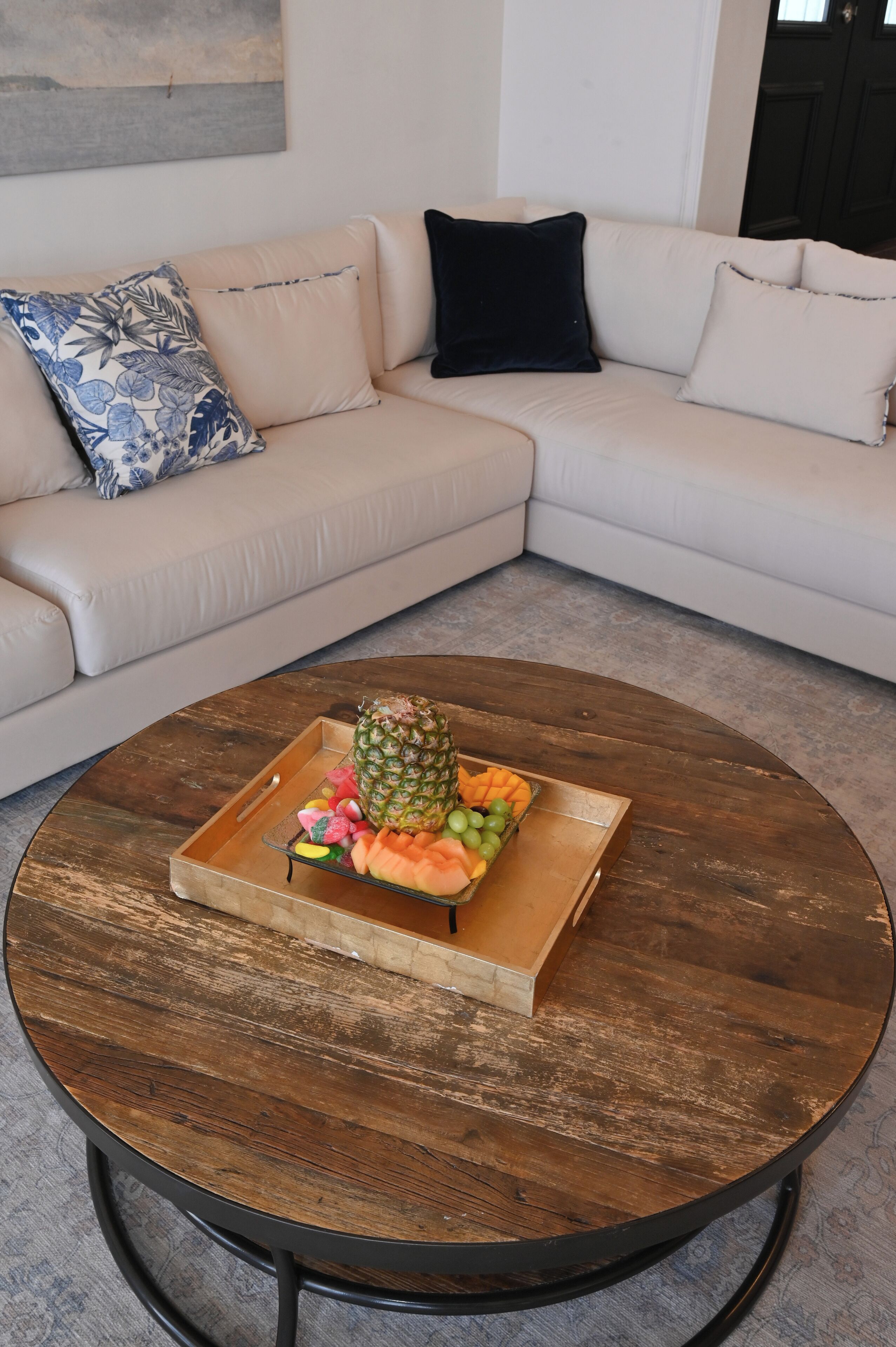 A round wooden coffee table with a tray of fruits and a pineapple, placed on a beige sofa with patterned pillows.