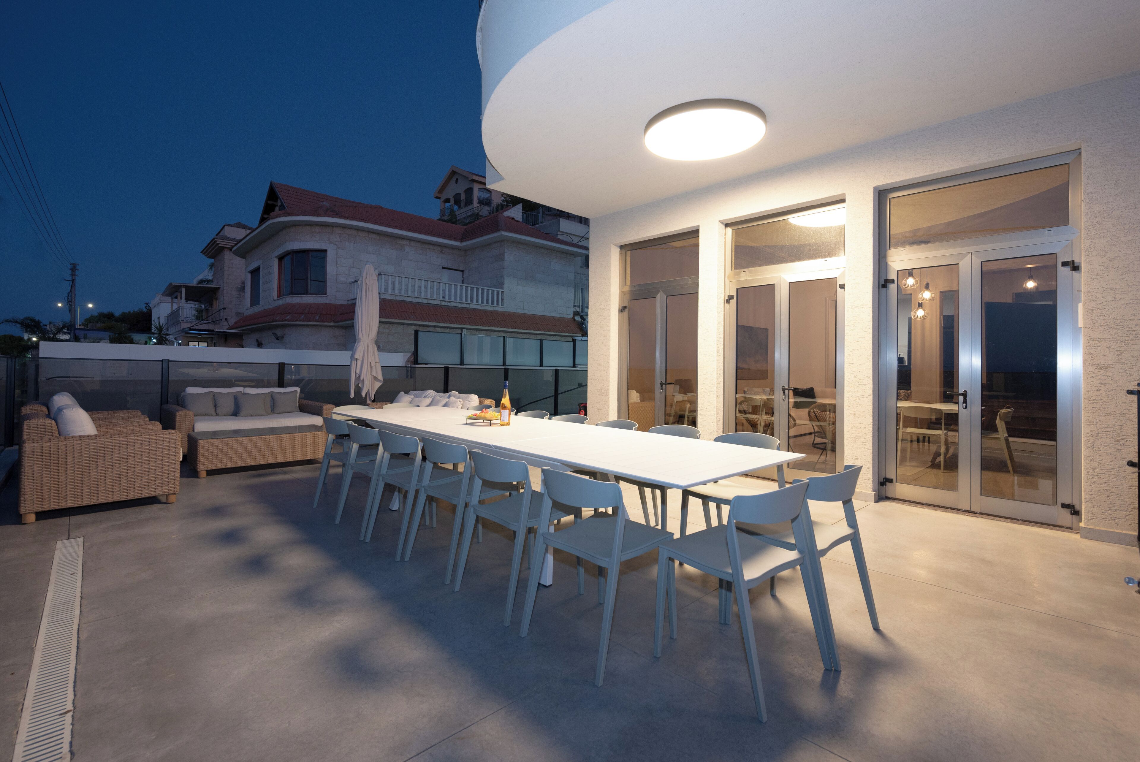 An outdoor dining area with a long white table and chairs, a wicker sofa, and a covered table with umbrellas.