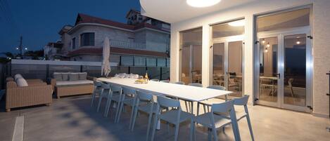 An outdoor dining area with a long white table and chairs, a wicker sofa, and a covered table with umbrellas.