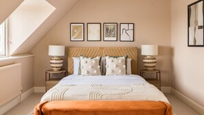 A bedroom with a sloped ceiling, a bed with white bedding and decorative pillows, two bedside tables with lamps, and four framed artworks on the wall. - Stylish West Kensington Duplex (London)