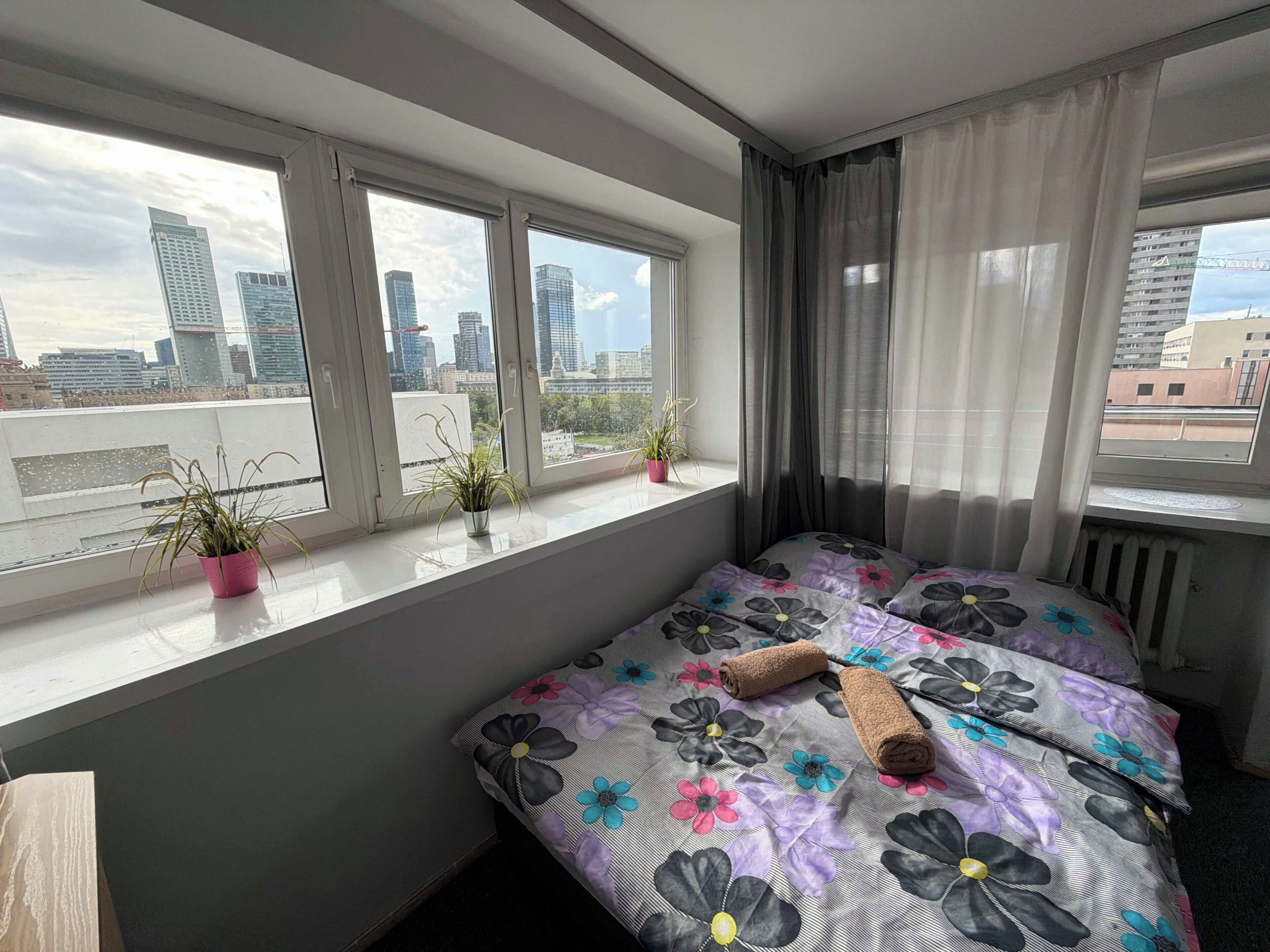 A bedroom with a bed, a window with curtains, and a view of a cityscape.