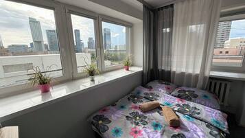A bedroom with a bed, a window with curtains, and a view of a cityscape.