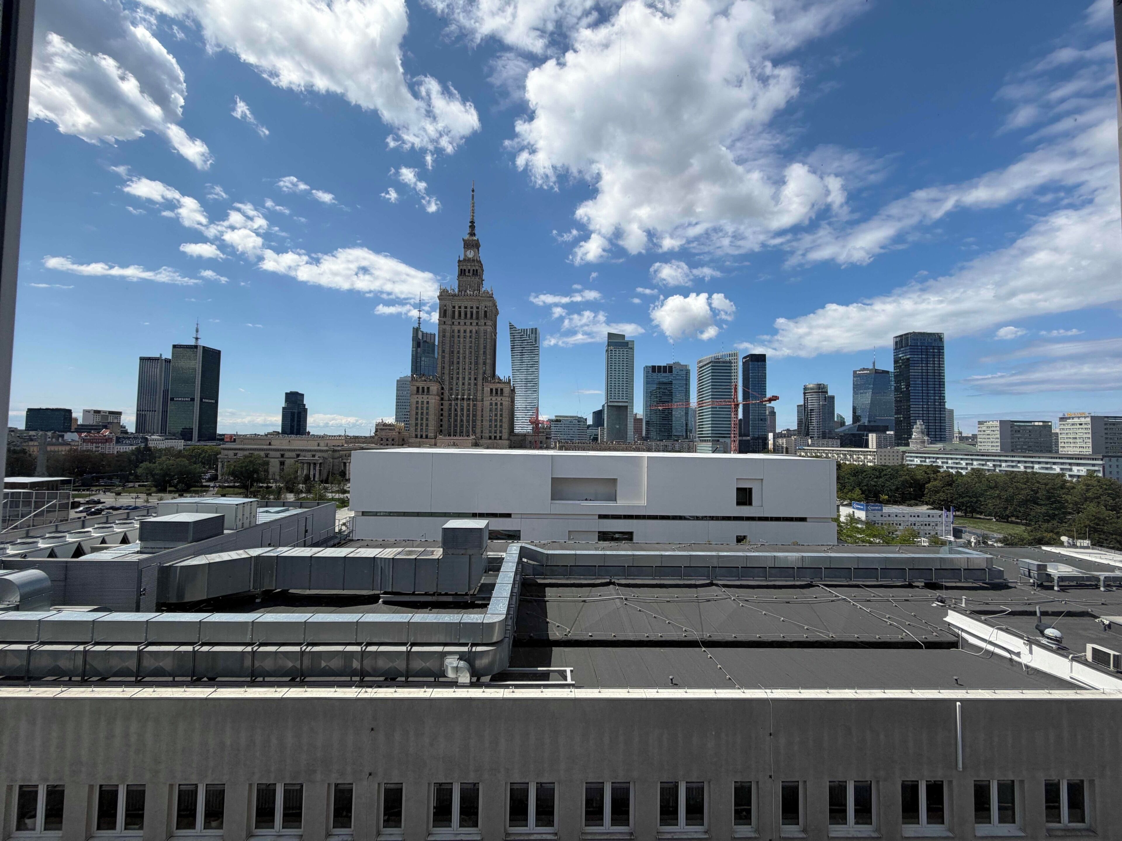 A cityscape with modern buildings, including a prominent tower with a spire, and a flat-roofed building in the foreground.