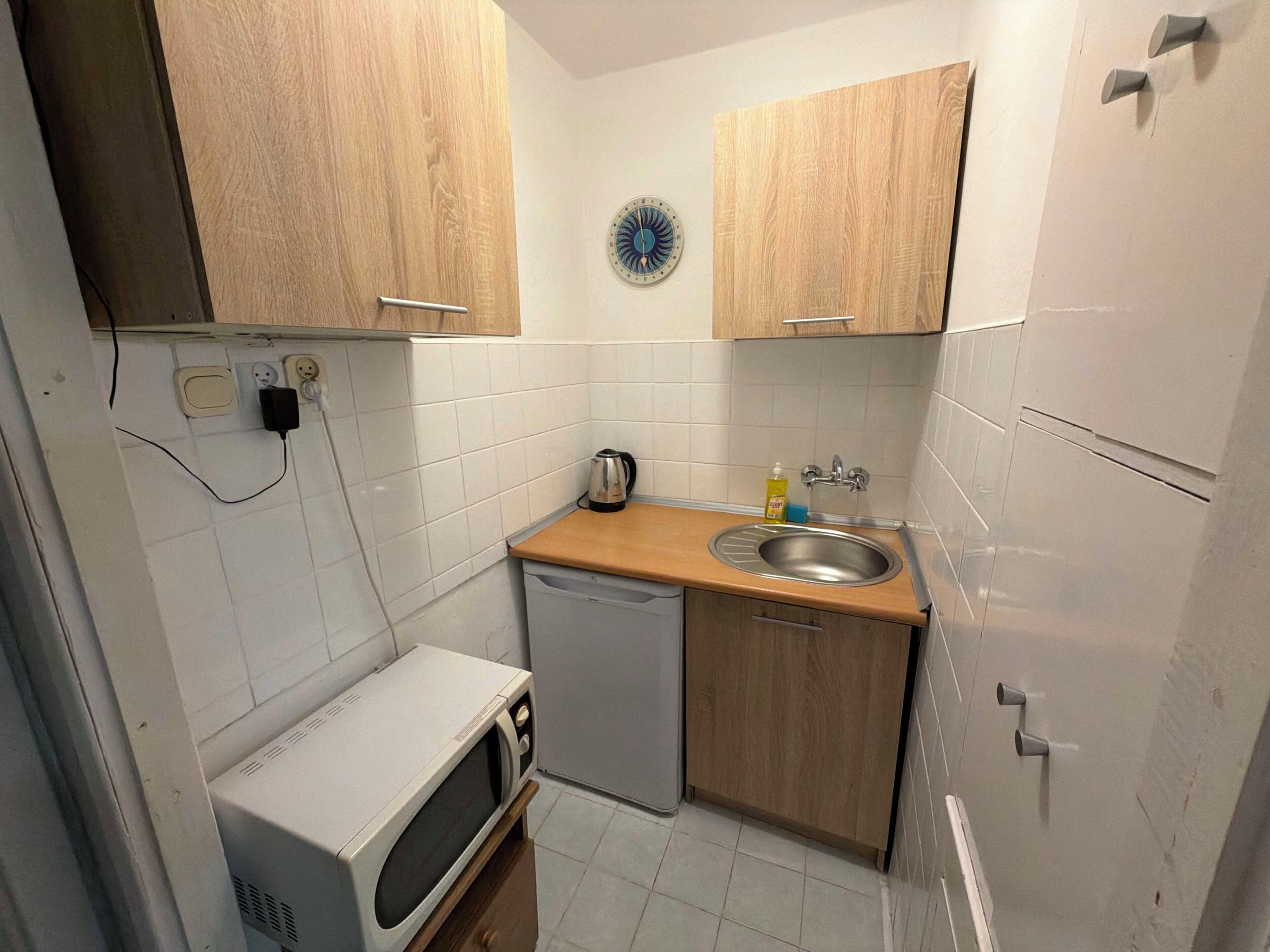 A compact kitchen with white tiles, a microwave, a sink, and wooden cabinets.