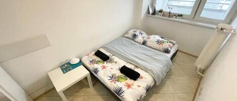 A small, well-lit bedroom with a bed, bedside table, and a window.