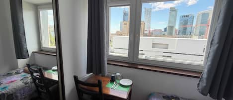 A room with a bed, a desk, and a chair. There is a window with a view of a cityscape.