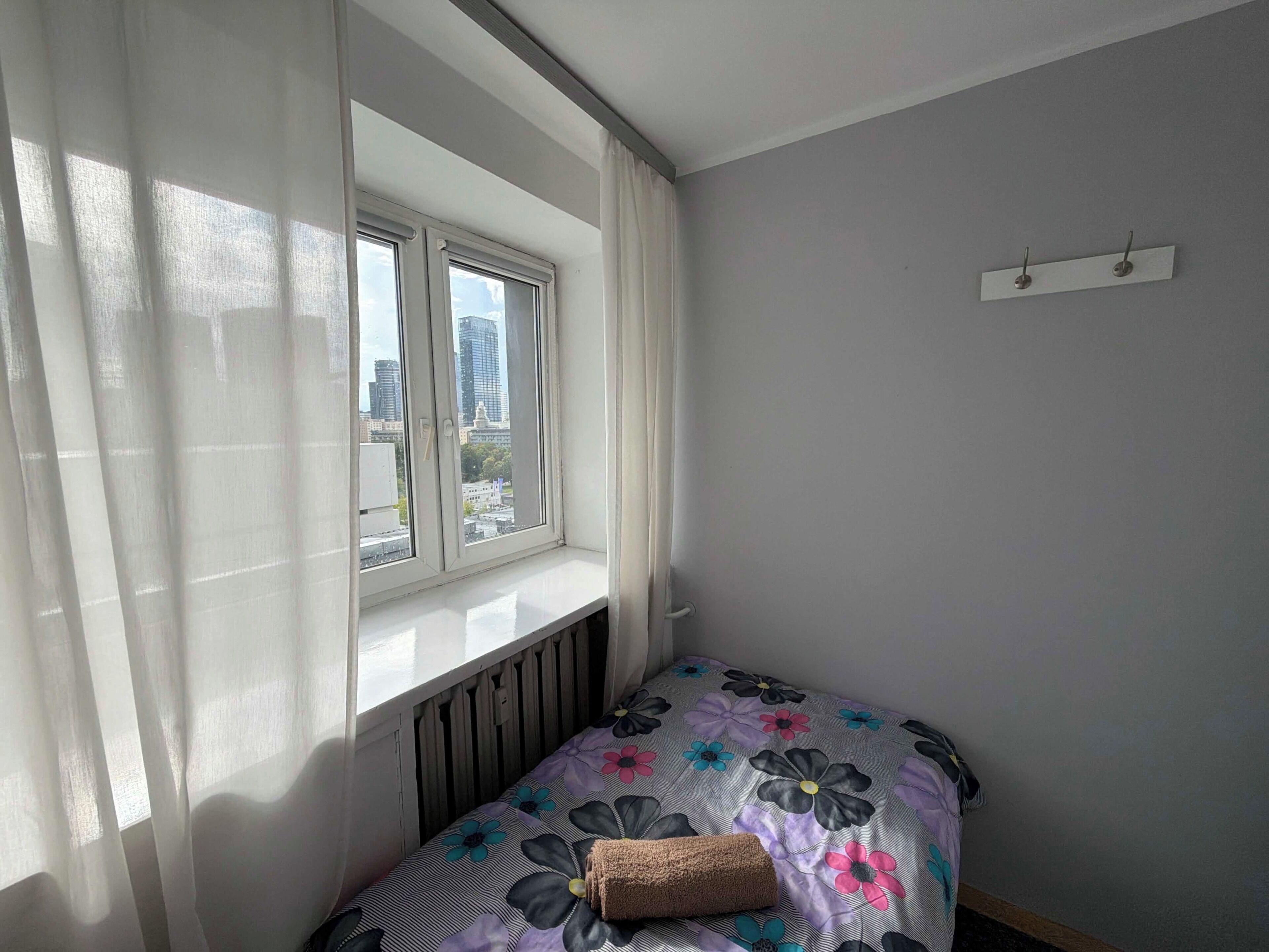 Een kleine kamer met een raam, een radiator, een bed met een bloemenbedekking en een wandplank.
