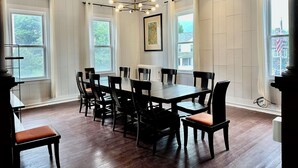 Een eetruimte met een grote tafel en stoelen, witte paneelde muren en meerdere ramen.