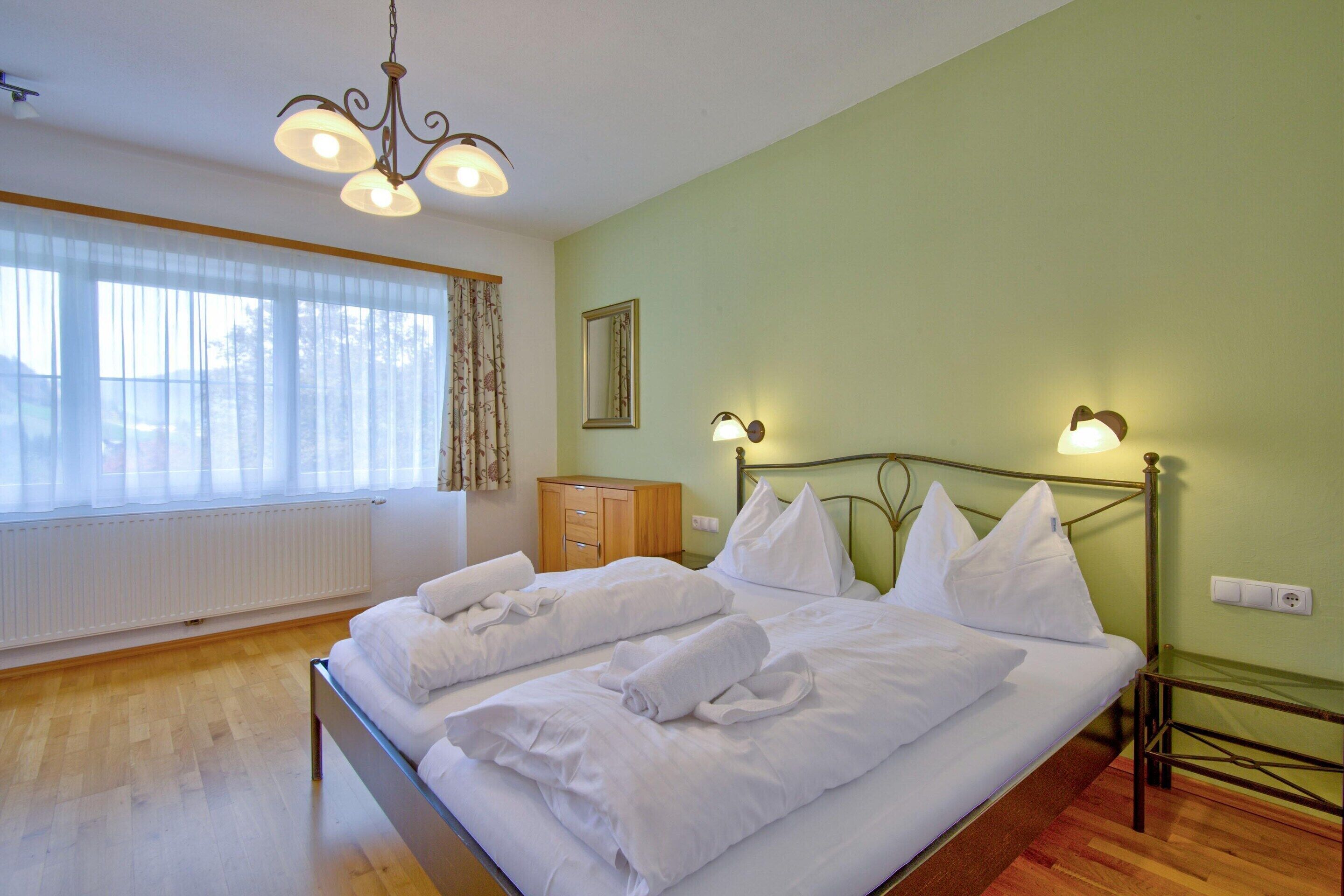 Ein ordentlich bezogenes Bett mit weißen Leinen und Kissen, ein Holzschrank, ein Spiegel an der Wand und eine Kristallleuchter.