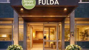 L’entrée d’un hôtel, avec une enseigne indiquant « Sleepy Hotels FULDA », présente un design moderne avec une porte d’entrée en verre, des marches menant à l’entrée et deux grands pots de plantes de chaque côté.
