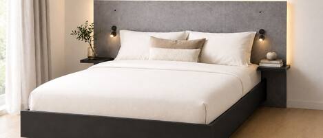 A modern bedroom with a large bed, dark headboard, bedside tables with lamps, and a plant on the nightstand.