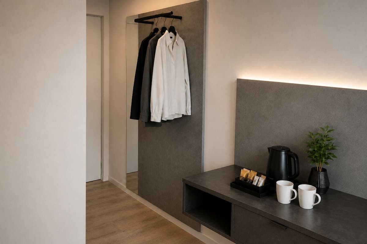 A modern hotel room with a wooden floor, a grey headboard, a bedside table with a black coffee maker, two white mugs, and a small potted plant.