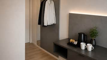 Una habitación de hotel moderna con piso de madera, cabecera gris, mesita de noche con cafetera negra, dos tazas blancas y una pequeña planta en maceta.