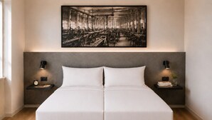 Un lit double avec une tête de lit grise, deux tables de chevet avec des lampes, une horloge et un tableau encadré au-dessus.