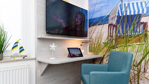 A modern living room with a TV mounted on a white wall, a small table with a tablet, a teal chair, and a beach-themed painting. - Komfortable Ferienwohnung mit Terrasse; Bensersiel (Esens)