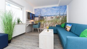 A modern living room with a blue sofa, a white coffee table, and a TV on a stand. There is a large window on one side and a painting of a beach scene on the wall. - Komfortable Ferienwohnung mit Terrasse; Bensersiel (Esens)