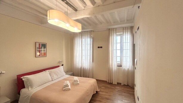 Ein Schlafzimmer mit Bett, Nachttisch, Pendelleuchte, Fenster mit Vorhängen und einem an der Wand hängenden Kunstwerk.