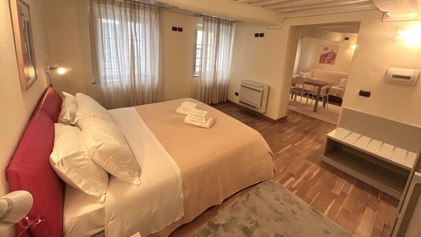 Un dormitorio con una cama grande, cabecera roja, un radiador y un área de comedor visible a través de una puerta abierta.