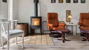 Een gezellige kamer met een witte bakstenen muur, een houtkachel, twee leren fauteuils, een kleine tafel en een witte stoel.