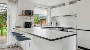 Een moderne keuken met een centraal eiland, witte kasten, een magnetron, een koffiezetapparaat en een spoelbak. Er staan barkrukken bij het eiland en een raam met uitzicht op groen.