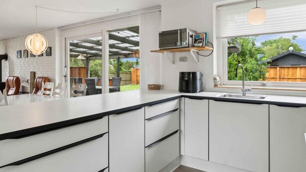 Een moderne keuken met witte kasten, een zwart aanrecht en een ingebouwde magnetron. Er is een eethoek met stoelen en een tafel zichtbaar door een open deur.