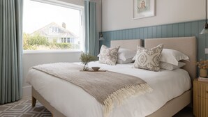 Una cama bien hecha con una manta y cojines color beige, una mesita de noche con lámpara, y una ventana con cortinas.