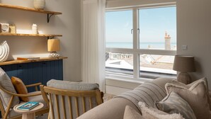 Una habitación acogedora con vistas al mar, que incluye una silla de madera, un sofá, una mesita con un libro y estantes con objetos decorativos.