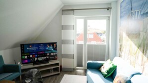 A living room with a flat-screen TV displaying a sports channel, a sofa, a fan, and a sliding glass door leading to a balcony. - Gemuetliche Ferienwohnung im Obergeschoss, Bensersiel (Esens)