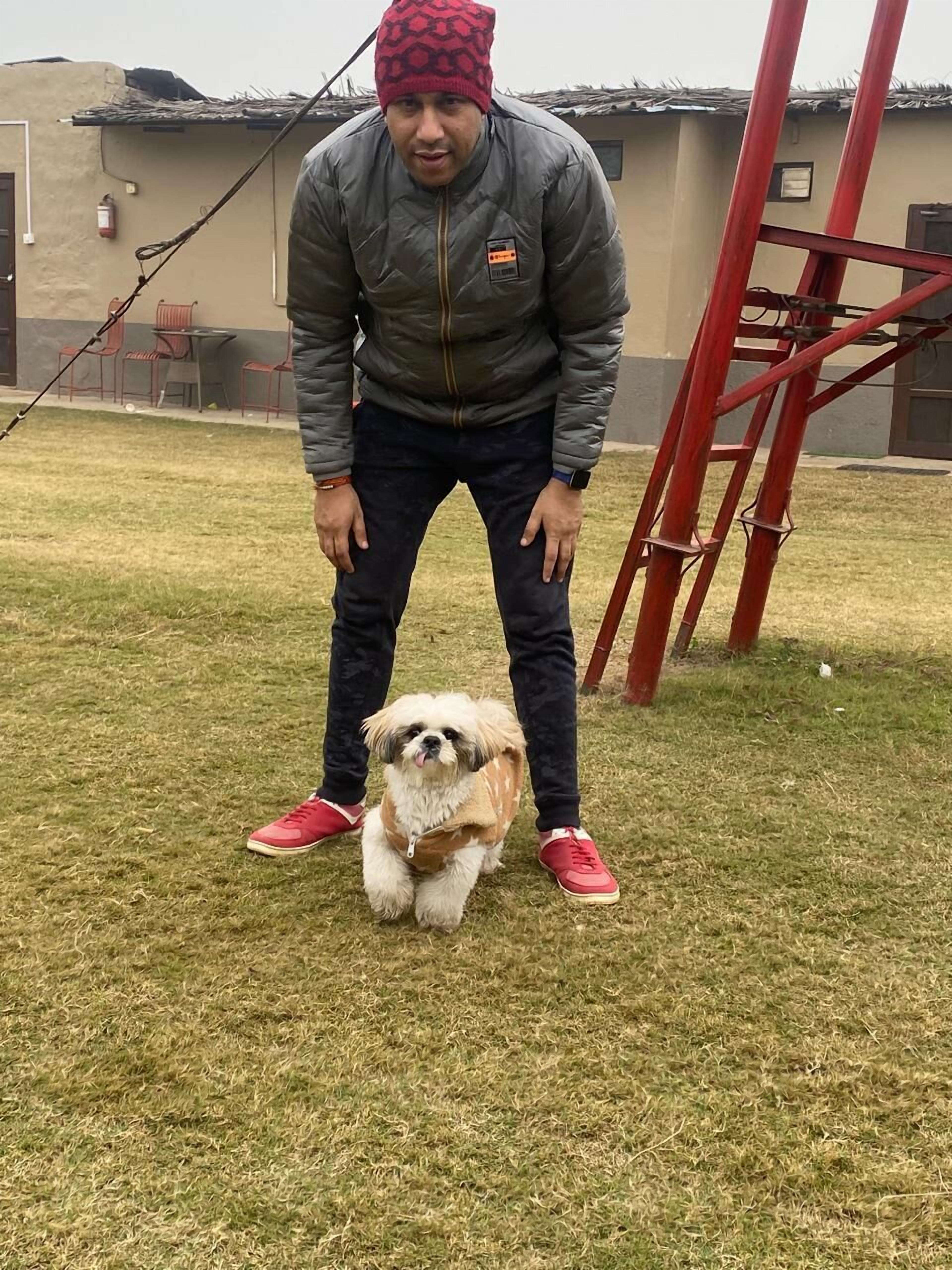 Una persona de pie sobre la hierba con un perro que lleva un chaleco, una estructura metálica roja y un edificio al fondo.