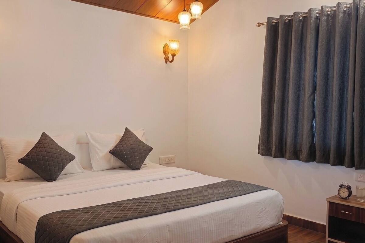 A bedroom with a wooden ceiling, a bed with a quilted bedspread, two wall-mounted light fixtures, a wall sconce, a window with curtains, and a bedside table with a clock.