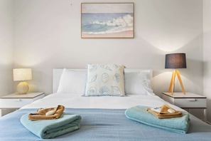 Une chambre d’hôtel avec un lit, un oreiller orné d’un motif de coquillage, une lampe et un tableau représentant une scène de plage.