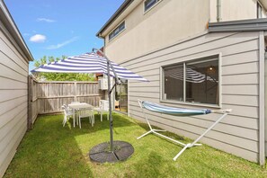 Ein Garten mit einer Terrasse, einem Tisch, Stühlen, einem gestreiften Sonnenschirm, einer Hängematte und einem Fenster.