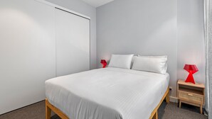 Un dormitorio con una cama, un armario, una mesita de noche con lámpara y una pequeña mesa auxiliar de madera.