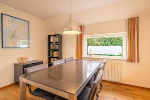 Un comedor con una mesa grande, sillas, una lámpara colgante, una estantería y una ventana con cortinas.