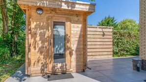 A wooden sauna structure with a glass door, a small porch, and a tiled patio area. - Villa With Finnish Sauna and Outdoor Shower (De Koog)