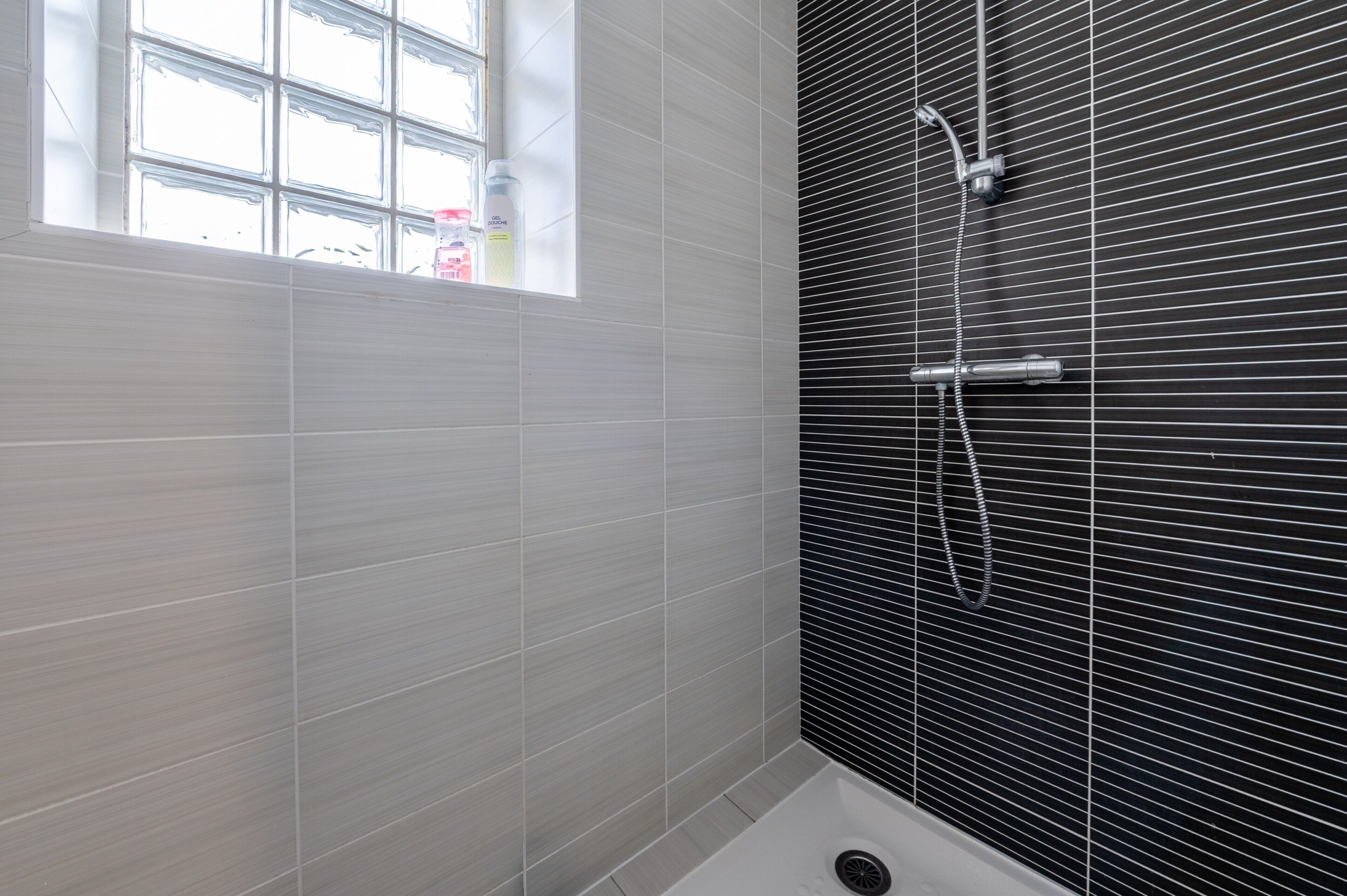 Une salle de bain avec des carreaux blancs, une fenêtre et une douche avec un mur recouvert de carreaux noirs.