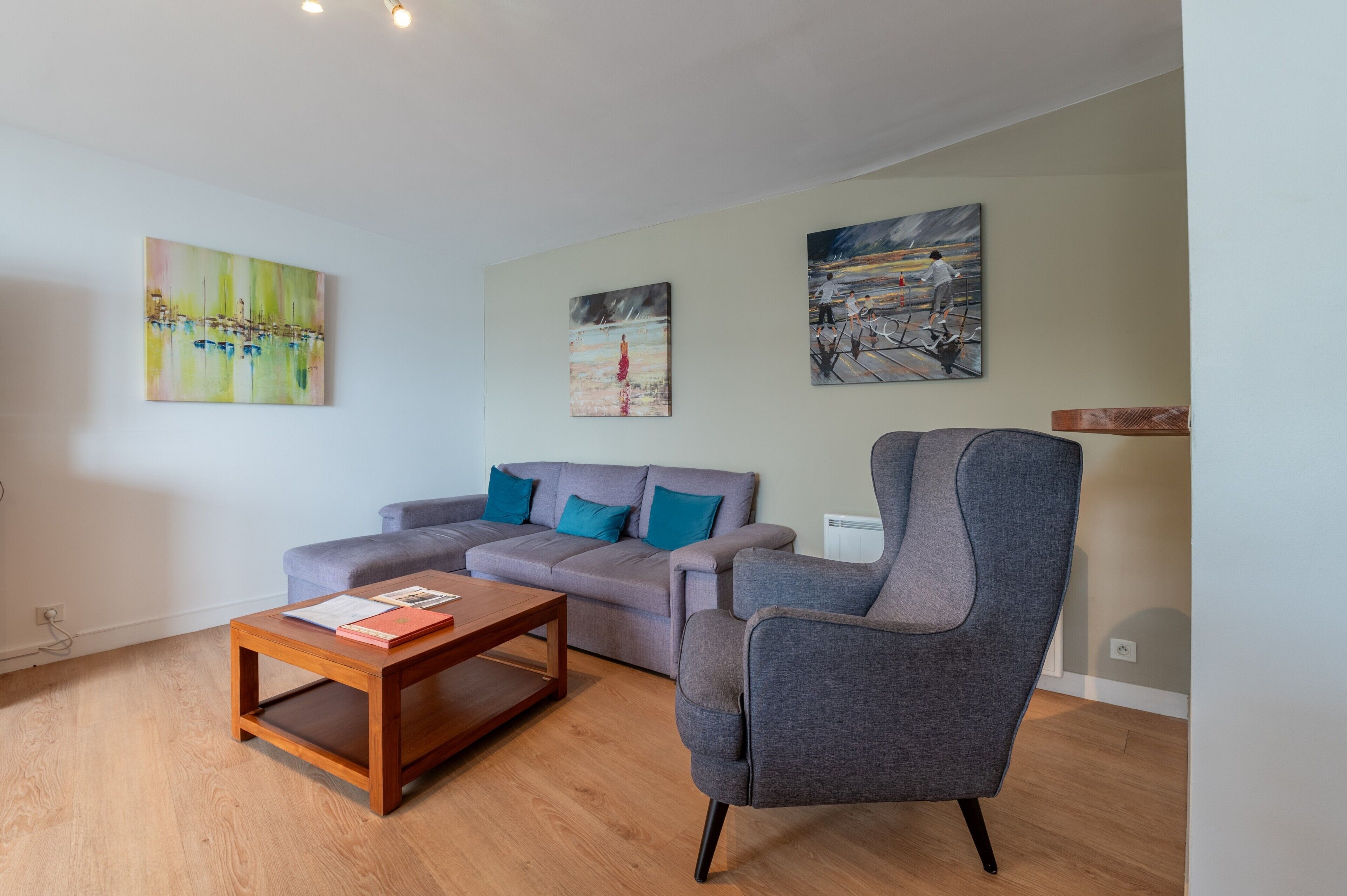 Un salon moderne avec un canapé gris, une table basse en bois et un fauteuil gris. Trois tableaux sont accrochés au mur.