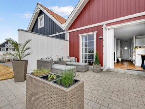 Et moderne hus med rødt facade, gråt tag og hvid dør. Der er udendørs møbler og plantekasser i forhaven.