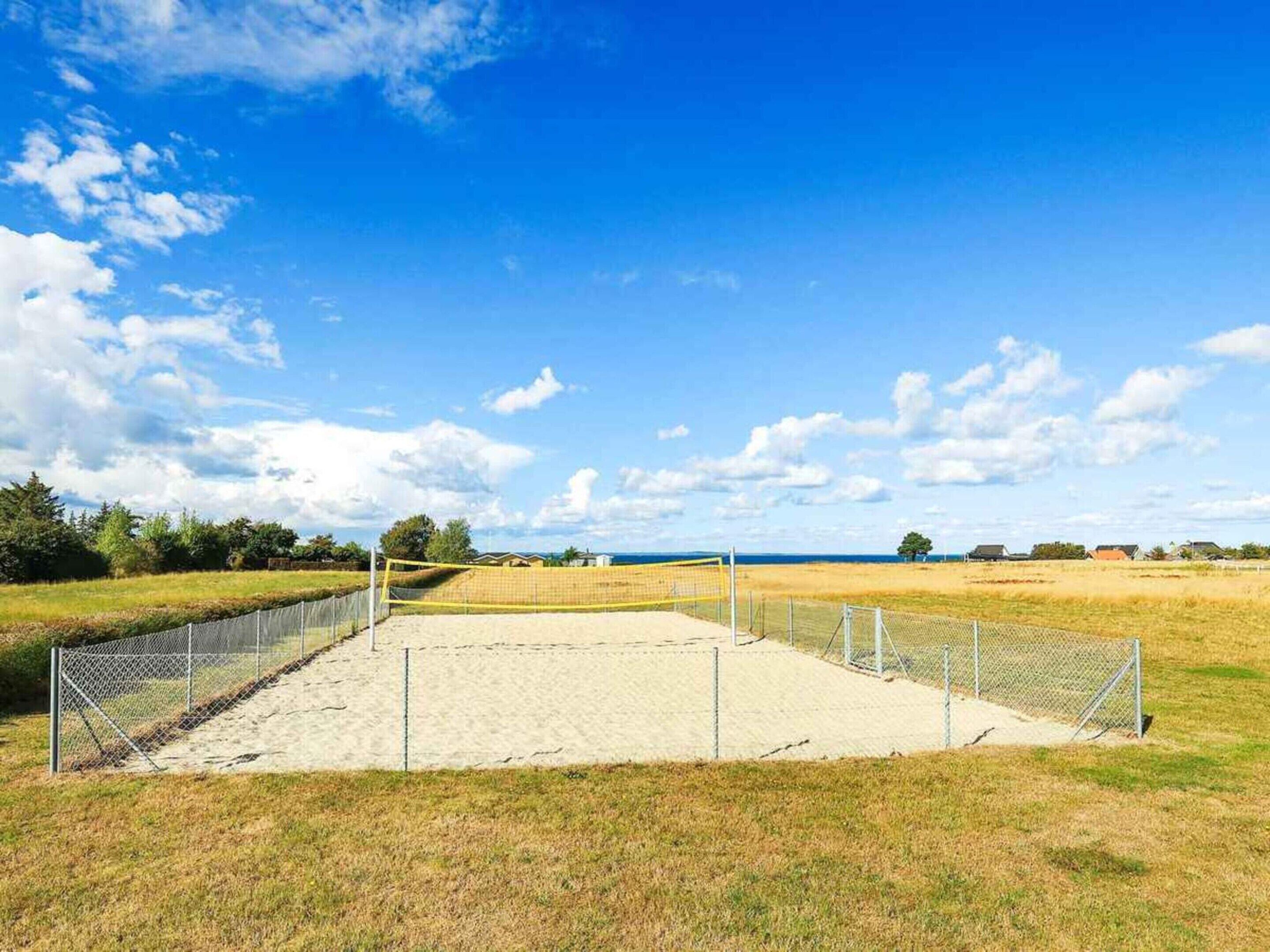Cancha de voleibol de arena con red, rodeada por una cerca de malla y zonas verdes.
