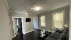 A spacious living room with a dark wood floor, a gray sofa, a coffee table, and a flat-screen TV mounted on the wall. - 2BR Apartment Near Wesleyan 102 (Middletown)