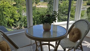 Ett runt glasbord med en krukväxt, två flätade stolar med kuddar och en utsikt över ett grönskande landskap genom stora fönster.
